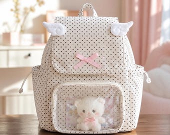 May include: A cream-colored backpack with a black polka dot pattern, featuring two white plush wings on the top flap, a pink ribbon bow, and a clear front pocket containing a small white plush bear with a pink bow around its neck.