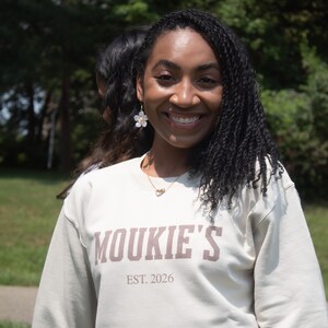 May include: A beige crewneck sweatshirt featuring the text MOUKIE'S and EST. 2026 printed in brown block lettering on the chest. The garment has long sleeves and a ribbed neckline, providing a simple and functional design for casual wear.