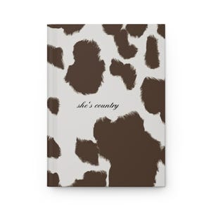 Può includere: Un diario con copertina rigida con un motivo a pelle di mucca marrone e bianco. Le parole "she's country" sono stampate in corsivo sulla copertina. Il diario è chiuso e ha un dorso bianco.