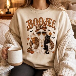 May include: A beige crewneck sweatshirt featuring a graphic of a ghost wearing a plaid bow, heart-shaped sunglasses, and a leopard print scarf. The ghost holds an iced coffee and a black cat, surrounded by pumpkins, bats, and leaves with the text BOOJEE.
