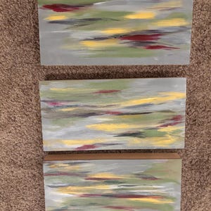 Könnte beinhalten: Set aus drei abstrakten Gemälden. Jedes rechteckige Stück hat einen grauen Hintergrund mit Pinselstrichen in Gelb, Grün, Rot und Weiß. Das Kunstwerk hat einen modernen, minimalistischen Stil, der sich für die Inneneinrichtung eignet.