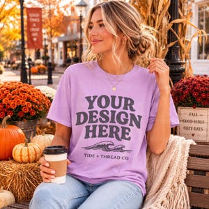 Op de afbeelding: Een lichtpaars t-shirt met korte mouwen voorzien van de donkergrijze tekst "YOUR DESIGN HERE" in een vet, golvend lettertype, gevolgd door een afbeelding van oceaangolven en de tekst "TIDE + THREAD CO" in een kleiner, strak schreefloos lettertype.