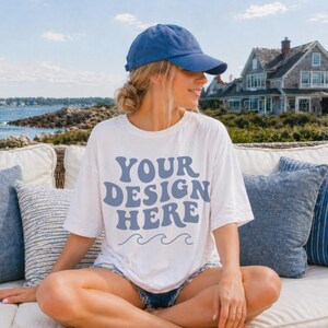 Puede incluir: Una camiseta blanca de manga corta con el texto azul "YOUR DESIGN HERE" sobre tres l&iacute;neas onduladas azules, combinada con una gorra de b&eacute;isbol azul lisa. La camiseta tiene un corte relajado y cuello redondo con un dise&ntilde;o gr&aacute;fico impreso.