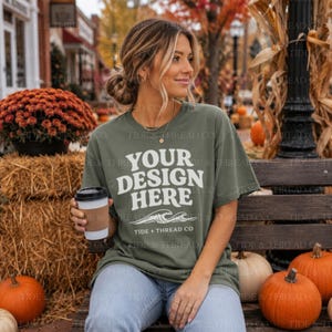 May include: An olive green short-sleeve t-shirt featuring the white text YOUR DESIGN HERE above a graphic of ocean waves and the text TIDE + THREAD CO. The shirt is worn with light blue denim jeans and is held with a disposable coffee cup.