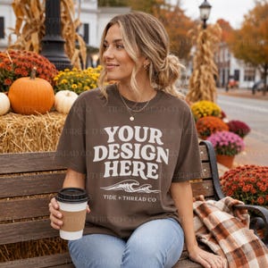 May include: A brown short-sleeve t-shirt featuring the white text YOUR DESIGN HERE above a graphic of ocean waves and the text TIDE + THREAD CO. The shirt is worn with light blue denim jeans and is held with a white paper coffee cup.