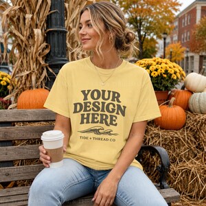 Op de afbeelding: Een lichtgeel T-shirt met korte mouwen en donkerbruine tekst met de tekst YOUR DESIGN HERE, een golfafbeelding en de tekst TIDE + THREAD CO eronder. Het shirt wordt gedragen met een lichtblauwe spijkerbroek en een gouden ketting.
