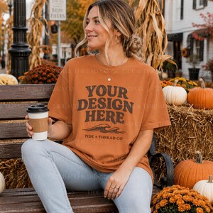 Op de afbeelding: Een oranjebruin t-shirt met korte mouwen en de donkerbruine tekst "YOUR DESIGN HERE" boven een golfafbeelding en de tekst "TIDE + THREAD CO". Het shirt wordt gecombineerd met een lichtblauwe spijkerbroek en een witte koffiebeker.