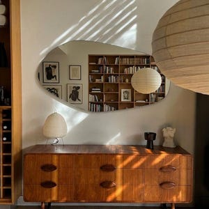 May include: A wooden mid-century style dresser with six drawers and oval handles. A lamp and a small sculpture are on top. A large, irregularly shaped mirror reflects a bookcase and artwork.