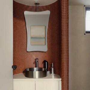 Könnte beinhalten: Ein modernes Badezimmer mit einem Waschbecken und einer Armatur aus Edelstahl. Die Wand hinter dem Waschbecken ist mit kleinen, quadratischen, rotbraunen Fliesen bedeckt. Über dem Waschbecken hängt ein großer, einzigartig geformter Spiegel.