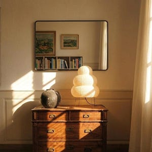 May include: A wooden dresser with four drawers, topped with a decorative lamp and a dark vase. A rectangular mirror hangs on the wall above, reflecting artwork and books. The lamp has a stacked, spherical design.