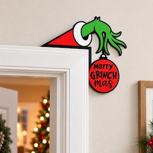 May include: A decorative door corner sign featuring a green hand emerging from a red and white Santa hat. The hand holds a round red Christmas ornament with the black text merry GRINCH mas printed on the front, designed for door frames.