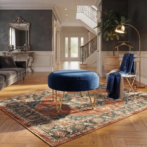 May include: A round, navy blue velvet ottoman with four thin, gold-toned metal hairpin legs. The ottoman is positioned in the centre of a room on a large, rectangular area rug featuring a traditional pattern in shades of rust, teal, and cream.