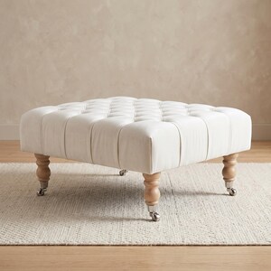 May include: A square white ottoman featuring a tufted top surface with button detailing. The piece is supported by four light-toned wooden legs, each finished with a silver-coloured metal caster wheel at the base for mobility and support.
