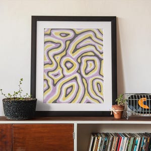Puede incluir: Impresión artística abstracta en un marco negro. La obra de arte presenta un patrón de formas arremolinadas en tonos lavanda, amarillo y gris. La impresión se exhibe en un mueble de madera con plantas y discos de vinilo.