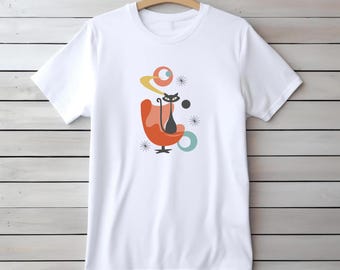 May include: White t-shirt featuring a mid-century modern design. The graphic includes a black cat sitting in an orange chair, with abstract shapes in yellow, teal, and black. The design has a retro, atomic age aesthetic.