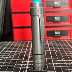 May include: A cylindrical gray tool with a matte finish, featuring a bright turquoise push-button at the top and a vertical pocket clip on the side. The lower section has three horizontal grooves, and a thin black ring separates the body segments.