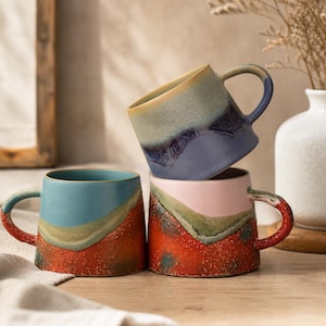 Puede incluir: Tres tazas de cerámica con un esmalte texturizado multicolor. Una taza está apilada sobre otras dos. Las tazas presentan combinaciones de colores en capas de azul, verde, naranja y rosa con un acabado moteado y asas curvas.