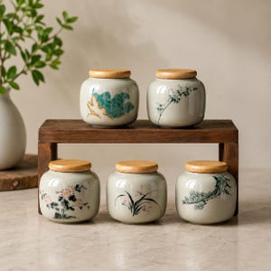 May include: Five small, round, light gray ceramic jars with wooden lids, arranged on a wooden stand. Each jar features a unique hand-painted design, including green mountains, bamboo stalks, pink flowers, thin grass-like plants, and pine branches.