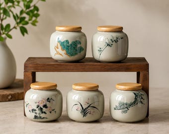 May include: Five small, light grey ceramic storage jars with round wooden lids. Each jar features a unique hand-painted botanical or landscape design, including flowers, bamboo, and pine branches, arranged on a small wooden display stand.