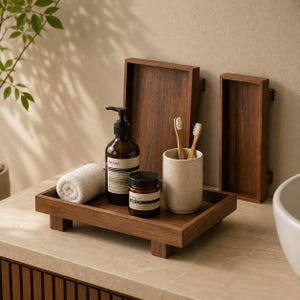 May include: A dark wood rectangular tray with four small feet holds a rolled white towel, a brown glass pump bottle labeled Aesop, a small brown jar labeled Parsley, and a speckled beige cup containing two bamboo toothbrushes.