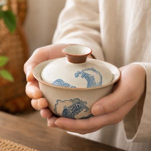 Może przedstawiać: Beżowa ceramiczna filiżanka do herbaty z pokrywką, zdobiona niebieskimi wzorami fal. Pokrywka ma mały brązowy uchwyt. Filiżanka trzymana jest w obu dłoniach, co podkreśla jej design. Tradycyjny element zestawu do herbaty.