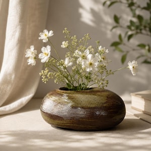 May include: A low, rounded ceramic vase with a mottled brown and olive green glaze. The vessel features a wide, circular opening at the top, currently holding a bouquet of delicate white flowers with thin green stems and small green buds.