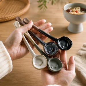 Puede incluir: Un juego de cuatro cucharas de cerámica con cuencos redondeados y mangos largos, que presentan una variedad de tonos tierra que incluyen crema, verde salvia, azul grisáceo moteado y negro sólido, con un acabado esmaltado y forma orgánica.