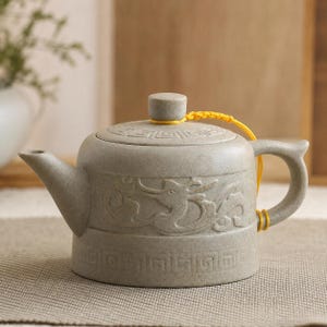 May include: A gray ceramic teapot with a dragon relief design. The teapot has a curved spout, a handle, and a lid with a small knob. A yellow cord is attached to the lid and handle. The teapot is on a neutral surface.