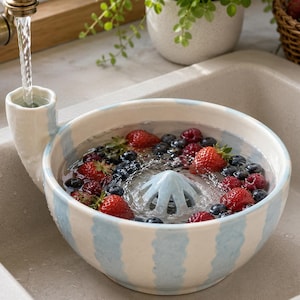 Puede incluir: Un cuenco de cerámica redondo con rayas verticales azul claro y un pico vertedor integrado, lleno de agua, fresas rojas frescas y arándanos azul oscuro. Un colador central en forma de cono se sitúa dentro del cuenco bajo un grifo.