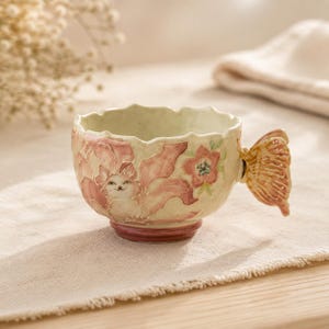 Puede incluir: Una taza de cerámica con borde festoneado, que presenta una ilustración pintada a mano de un gato rodeado de motivos florales rosas. La taza incluye un asa tridimensional con forma de mariposa en tonos naranja y marrón.