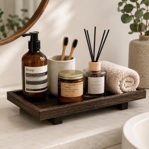 May include: A dark brown wooden tray holding an amber glass Aesop hand wash bottle, a ceramic cup with two bamboo toothbrushes, a small amber glass candle jar labeled Teakwood & Tobacco, a reed diffuser, and a rolled beige cotton washcloth.