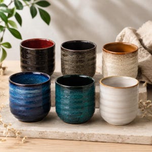 Könnte beinhalten: Ein Set aus sechs Keramikbechern in zylindrischer Form mit horizontaler Rippenstruktur. Die Becher haben verschiedene Glasuroberflächen, darunter tiefblau, blaugrün, weiß, dunkelbraun, gesprenkeltes Beige und ein schwarzer Becher mit rotem Rand.