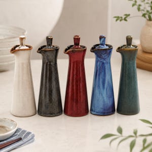 Può includere: Cinque dosatori per olio in ceramica con silhouette affusolata e coperchio abbinato. Il set include i colori bianco, grigio antracite, rosso intenso, blu vivace e verde ottanio, ciascuno con finitura lucida e un piccolo beccuccio versatore.