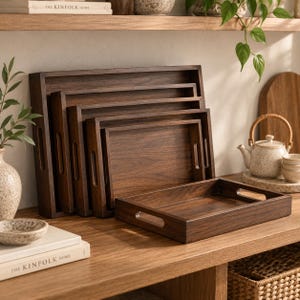 May include: A set of rectangular dark brown wooden serving trays with cutout handles, nested together in a graduated arrangement from largest to smallest, with one tray placed horizontally in the foreground on a wooden surface.