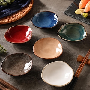 May include: A set of six small, round ceramic dipping bowls with irregular, organic edges. The bowls feature a variety of glossy, reactive glazes in solid red, deep blue, dark green, beige, speckled grey, and off-white, each with a dark rim.