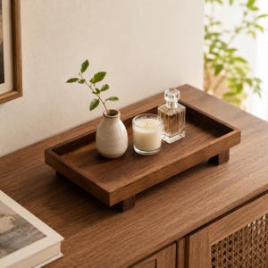 May include: A rectangular wooden tray with four short feet, holding a small speckled ceramic vase with a green leafy sprig, a white candle in a clear glass votive, and a clear glass perfume bottle with a faceted stopper on a wooden surface.