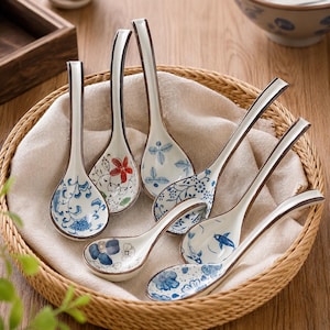Könnte beinhalten: Ein Set aus sieben Keramik-Suppenlöffeln mit langen Stielen und tiefen Laffen, die jeweils mit handbemalten blau-weißen Blumen-, Pflanzen- oder Fischmotiven verziert sind, wobei ein Löffel einen einzelnen roten Blumenakzent aufweist.