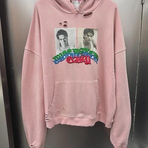 May include: A light pink hooded sweatshirt featuring distressed detailing around the neckline, cuffs, and hem. The front displays two black and white portraits of a person above colorful, stylized text that reads "MISMATCHED GARY".