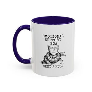Puede incluir: Una taza de cerámica blanca con asa e interior azul oscuro. El frente presenta una ilustración en blanco y negro de una persona con una boa constrictora alrededor del cuello, con el texto "EMOTIONAL SUPPORT BOA" y "NEED A HUG?"