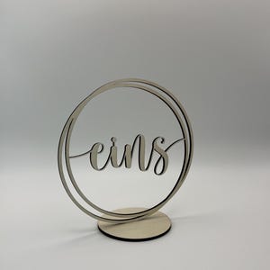 Könnte beinhalten: Eine Tischkarte aus Holz mit dem Wort "eins" in Schreibschrift, eingefasst in einem doppelten Kreis. Die Karte steht auf einem kleinen, runden Holzsockel. Das Design ist schlicht und elegant.