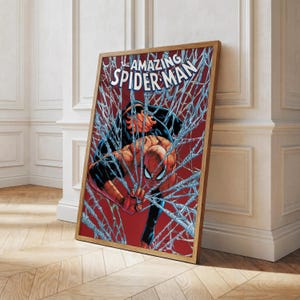 K&ouml;nnte beinhalten: Gerahmter Comic-Kunstdruck mit Spider-Man in einer dynamischen Pose, umgeben von seinem Netz. Der Titel "The Amazing Spider-Man" ist oben zu sehen. Das Kunstwerk hat einen roten Hintergrund und einen Holzrahmen.