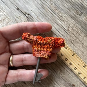 May include: A small, orange and red knitted sweater is placed on a silver key. The key is about 3 inches long and the sweater is about 1 inch wide. The key and sweater are sitting on a wooden surface.