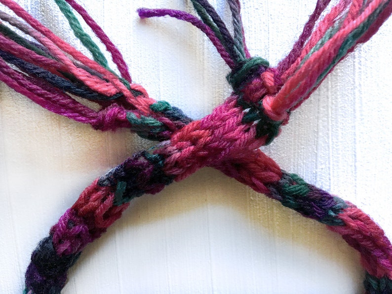 May include: A close-up of a multi-colored braided cord with pink, green, and purple yarn. The cord is tied in a knot.
