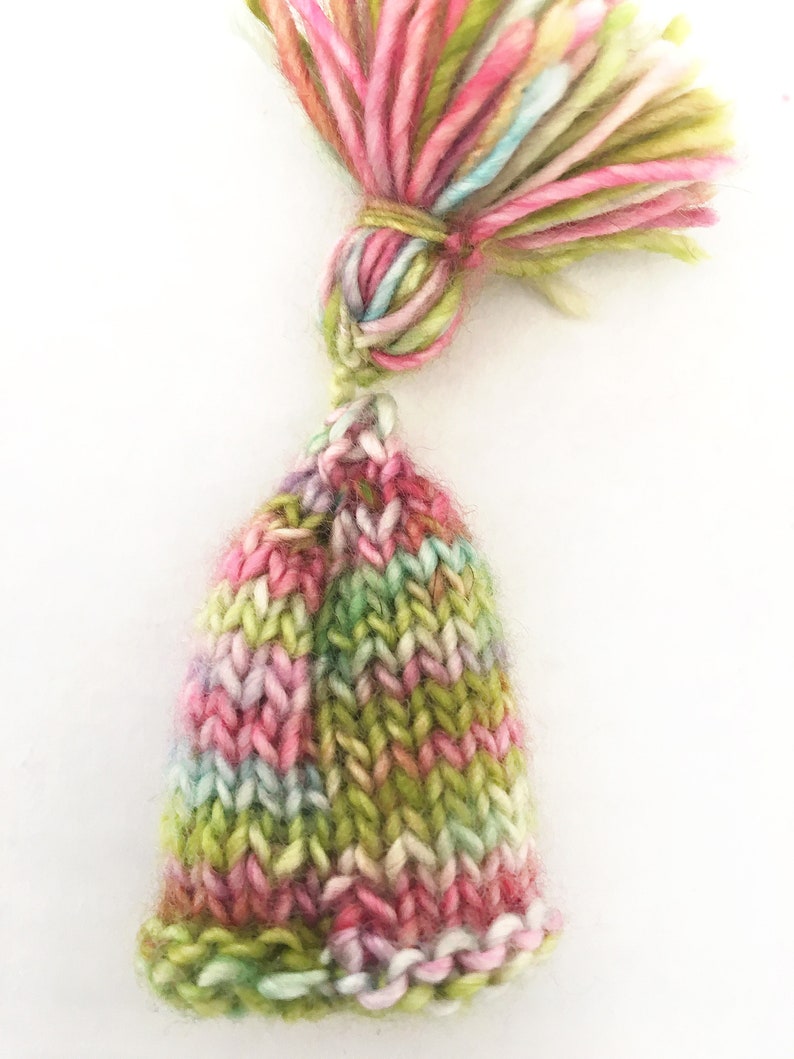 May include: A small, knitted hat with a green, pink, and white striped pattern. The hat has a matching tassel on top.