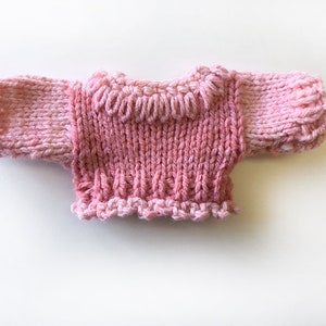 May include: A pink knitted sweater with a ruffled neckline and long sleeves.