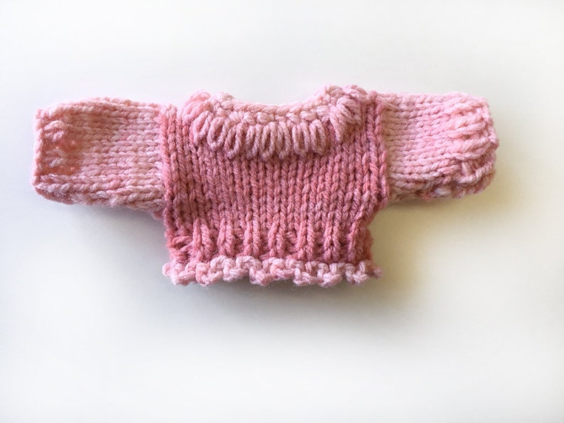 May include: A pink knitted sweater with a ruffled neckline and long sleeves.