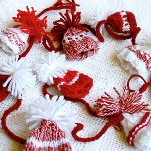 Puede incluir: Una guirnalda de gorros de punto rojos y blancos con pompones blancos. Los gorros est&aacute;n conectados por hilo rojo y blanco.