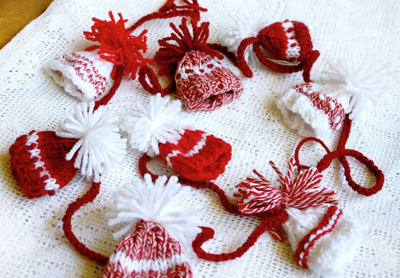 Puede incluir: Una guirnalda de gorros de punto rojos y blancos con pompones blancos. Los gorros est&aacute;n conectados por hilo rojo y blanco.