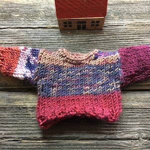 May include: A small, hand-knitted sweater in shades of red, pink, blue, and brown. The sweater has a striped pattern and a crew neck.