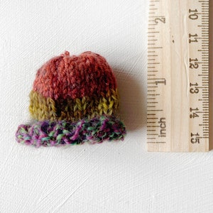 Puede incluir: Un pequeño gorro tejido a mano con un esquema de color rojo, verde y morado. El gorro tiene un diámetro de aproximadamente 5 cm.